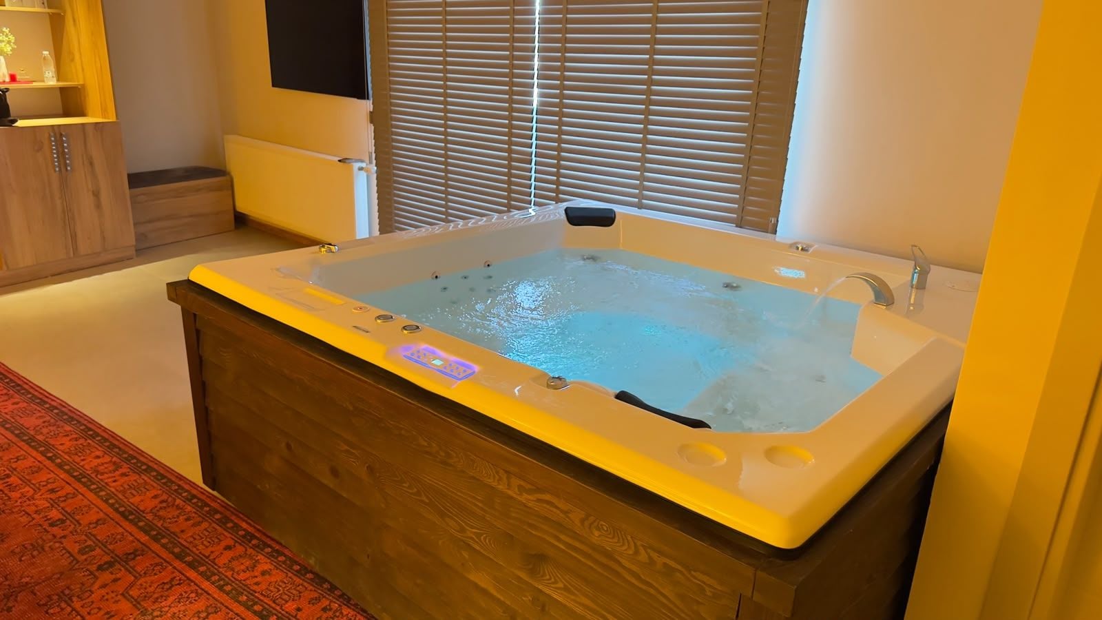 Un jacuzzi en una habitación con un armario de madera, una ventana con persianas y una alfombra estampada.