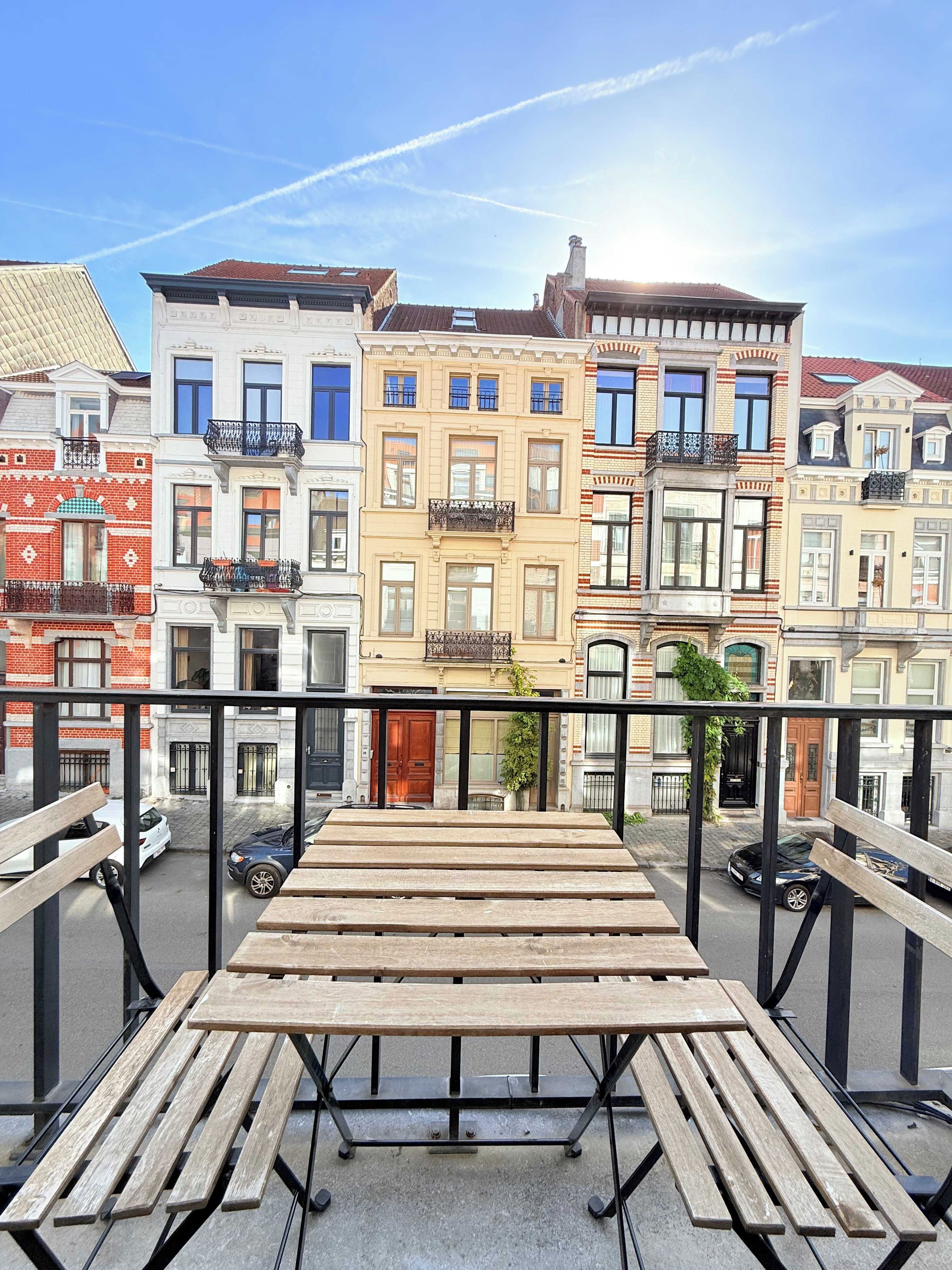 A balcony with a wooden table and benches, overlooking a row of colorful buildings with balconies.