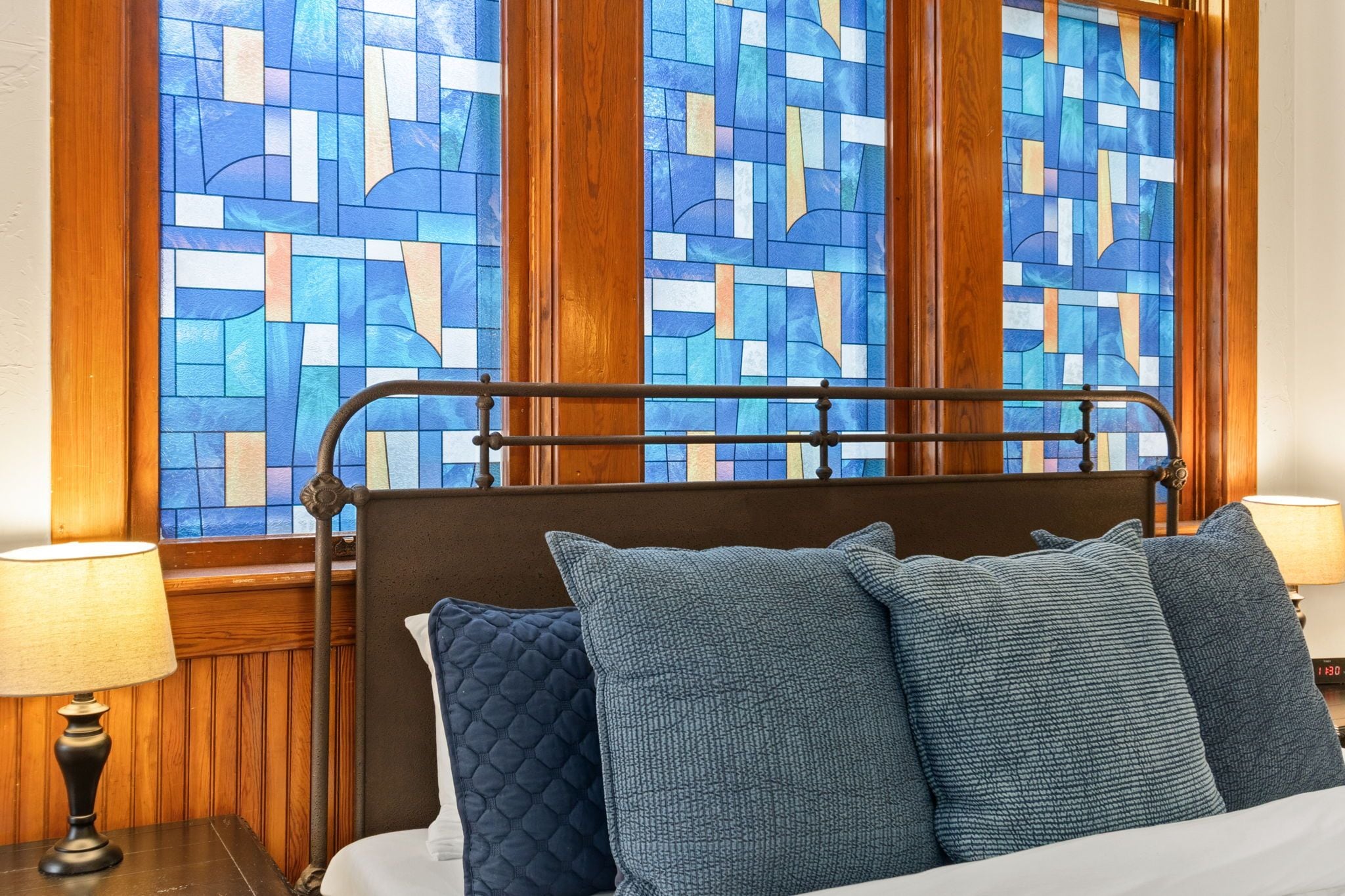 A bed with a headboard, two pillows, a bedside lamp, and a window with a stained glass design.