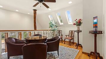 A spacious living room with a wooden floor, a ceiling fan, a chess table, leather chairs, a wooden coffee table, a floral rug, a potted plant, and a decorative vase.
