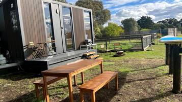 A modern wooden deck with a table and bench, a small outdoor kitchen area, and a fenced yard with trees.