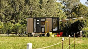 A small, modern cabin with a sliding door, situated in a grassy field with trees in the background.