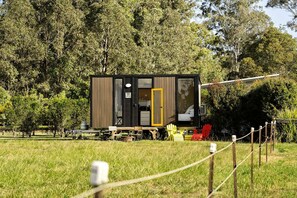 A small, modern cabin with a sliding door, situated in a grassy field with trees in the background.