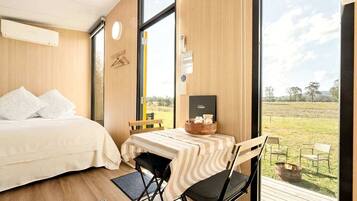 Une chambre à coucher avec un lit, un bureau avec une chaise et une vue sur un champ verdoyant, visible à travers de grandes fenêtres.
