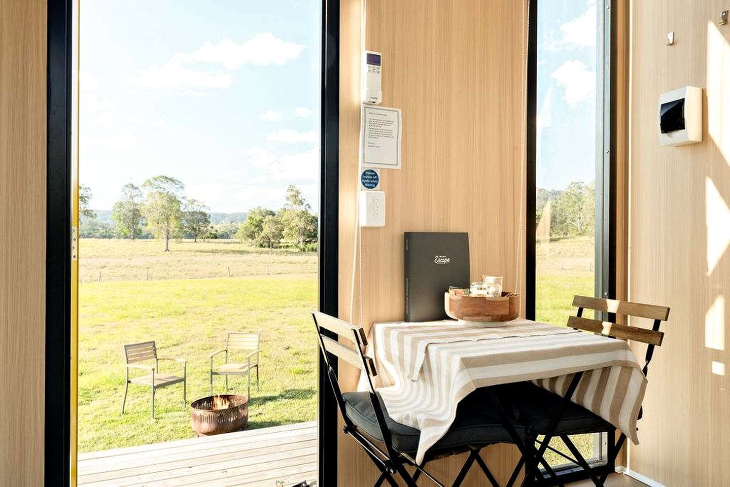Ein Zimmer mit einem großen Fenster, einem Tisch mit weißem Tuch, zwei Stühlen, einer kleinen Holzkommode und einer Metallschüssel auf dem Boden.
