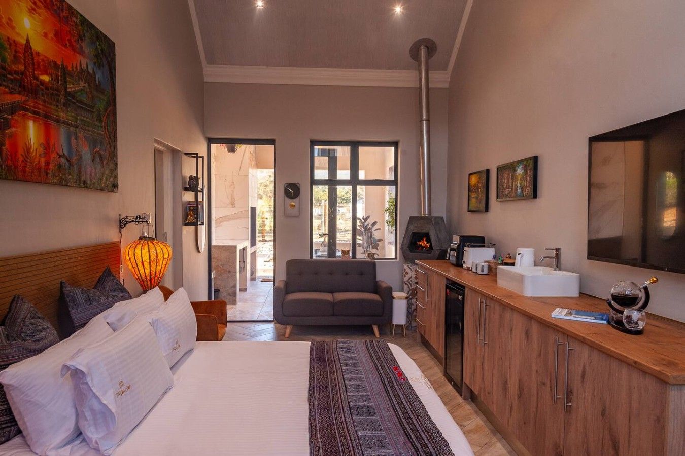A modern bedroom with a large bed, a sofa, a fireplace, and a kitchen area with a coffee machine.