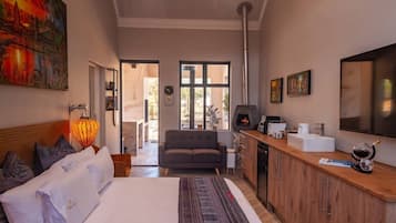 A modern bedroom with a large bed, a sofa, a fireplace, and a kitchen area with a coffee machine.