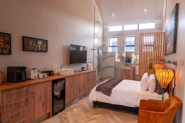 A modern hotel room with a bed, bedside table, a shower, a desk with a computer, and artwork on the walls.