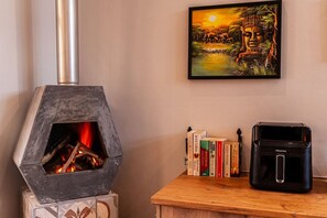 A room with a wood-burning stove, a framed painting, a stack of books, and a black coffee maker.