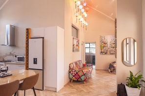A modern kitchen with white cabinetry, a dining table with chairs, a colorful sofa, and a hanging light fixture.