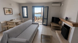 Un salon avec un canapé, une télévision, une cheminée et une vue sur la mer, visible à travers une porte ouverte.