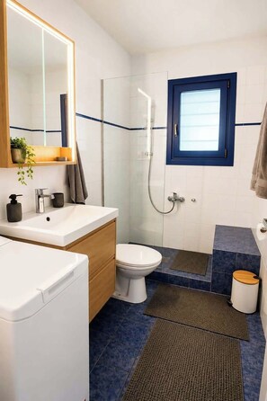 Une salle de bain moderne avec une douche à l’italienne en verre, des toilettes blanches, un lavabo avec un miroir et une fenêtre située au-dessus des toilettes.