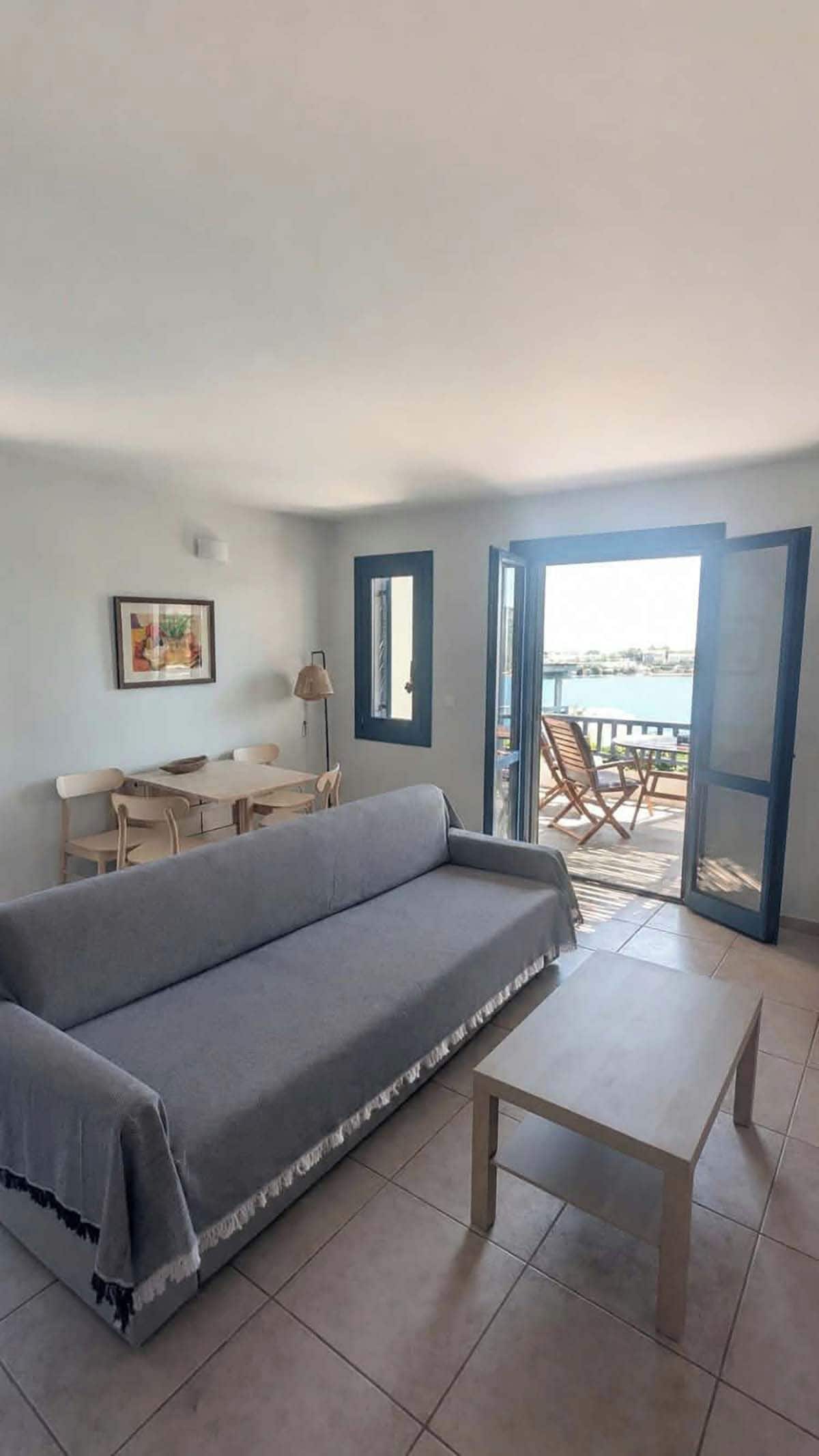 A living room with a sofa, a coffee table, a dining table with chairs, and a view of the sea through an open door.