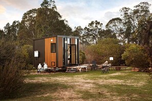 A modern, compact mobile home with a wooden exterior and large windows, situated in a grassy area with trees in the background.