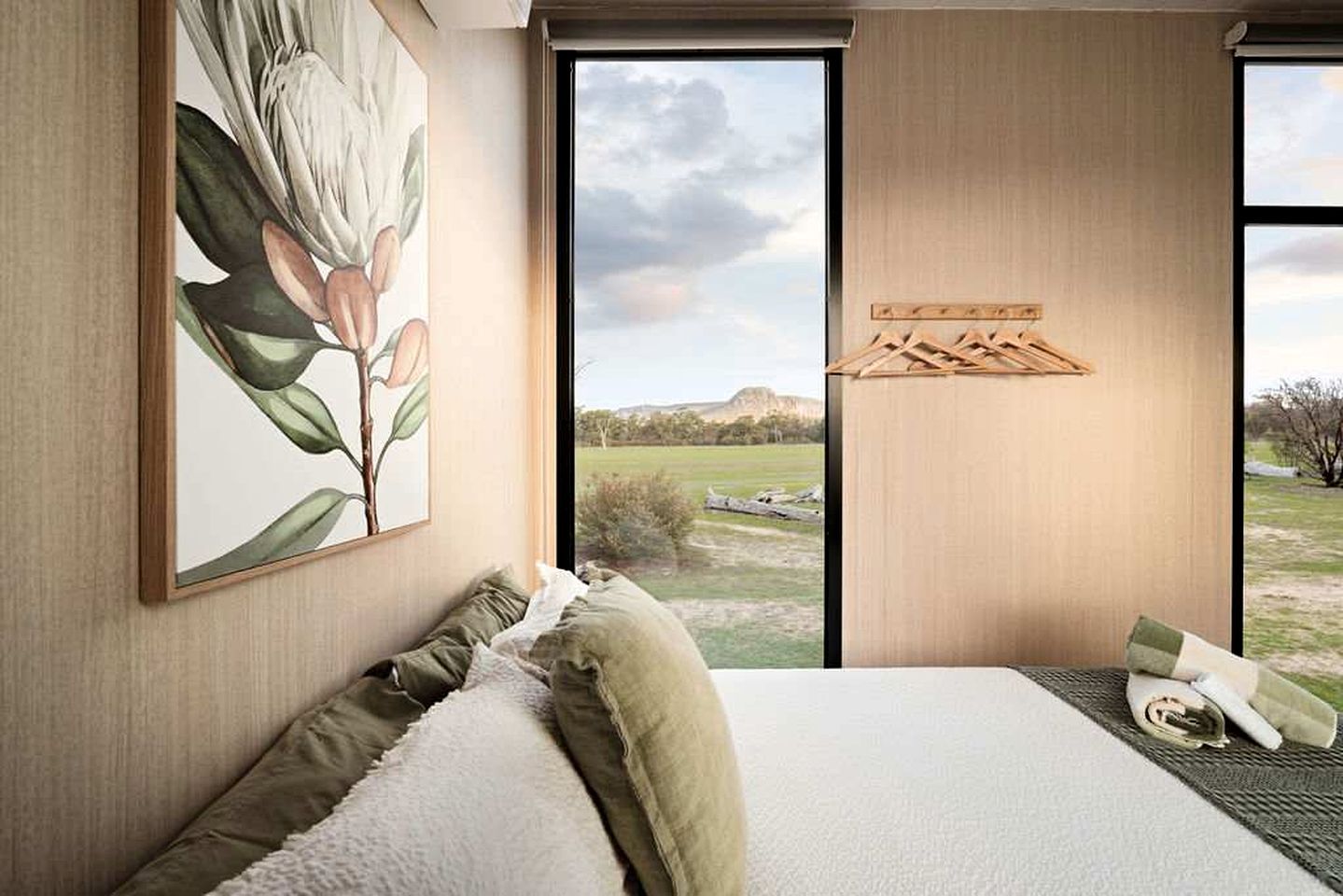 Ein Schlafzimmer mit großem Fenster, einem Bett mit Kissen, einem Bild an der Wand und einem Holzregal.