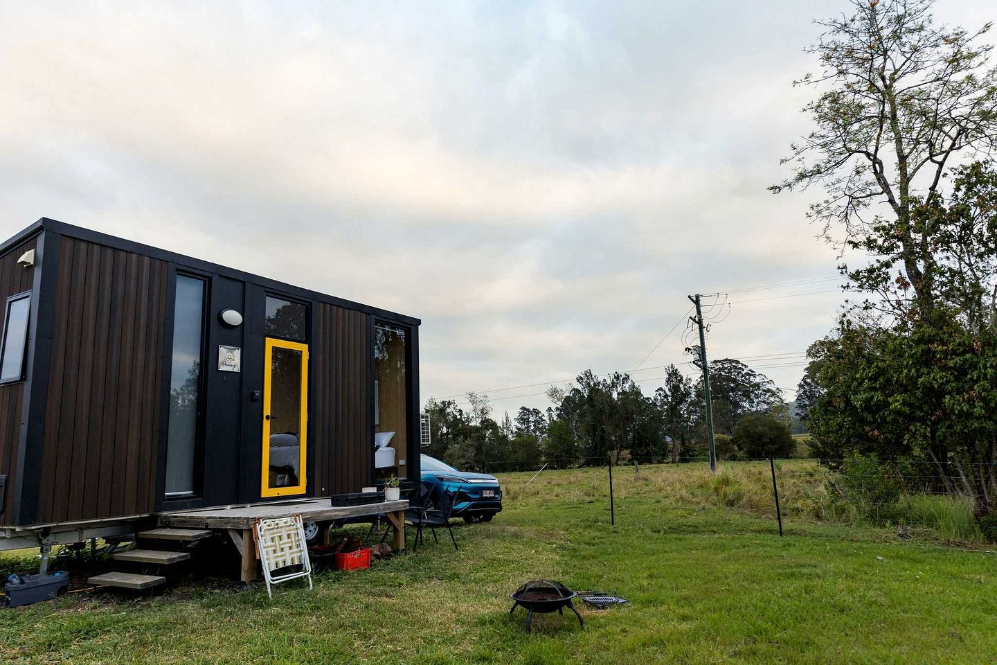 Una casa móvil moderna y compacta, con una puerta amarilla distintiva, ubicada en una zona con césped y un coche azul estacionado cerca.