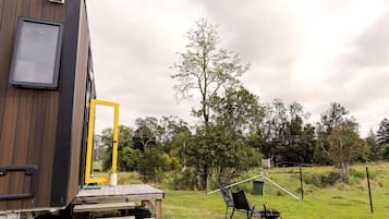Una cabaña de madera con puerta, un pequeño espacio exterior con sillas y una mesa, y un campo con árboles al fondo.
