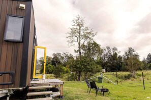 Una cabaña de madera con puerta, un peque ño espacio exterior con sillas y una mesa, y un campo con árboles al fondo.