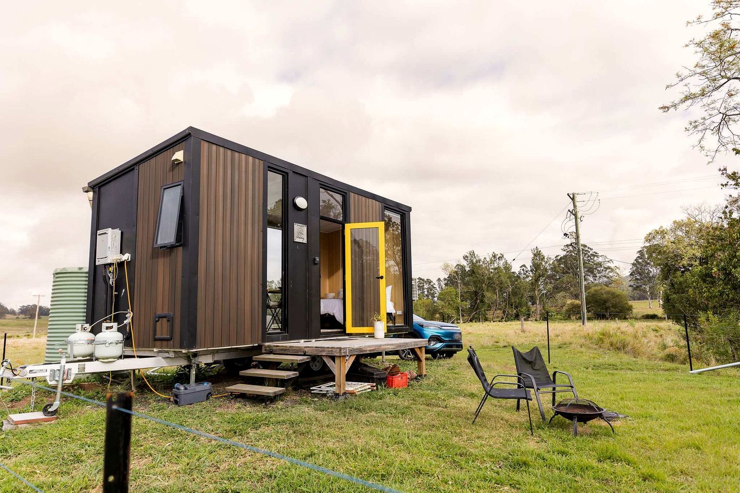 Una cabaña pequeña y moderna con fachada de madera y una puerta amarilla, ubicada en un campo cubierto de césped con un coche azul cerca.