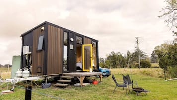 Una cabaña pequeña y moderna con fachada de madera y una puerta amarilla, ubicada en un campo cubierto de césped con un coche azul cerca.