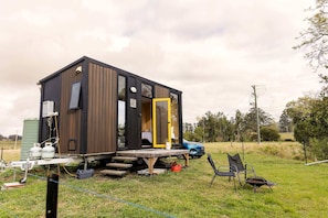 A small, modern cabin with a wooden exterior and a yellow door, situated on a grassy field with a blue car nearby.