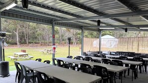 Uma área externa coberta para refeições, com mesas e cadeiras, uma lixeira e vista da paisagem ao redor.