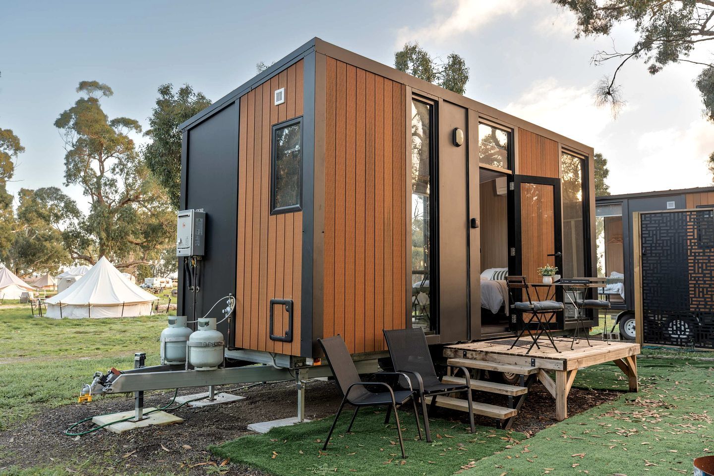 A modern, compact mobile home with a wooden exterior, a patio with outdoor furniture, and a visible propane tank.