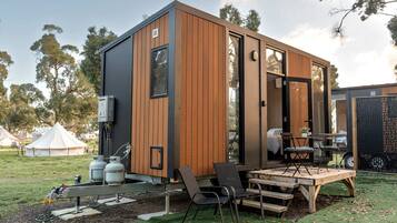 A modern, compact mobile home with a wooden exterior, a patio with outdoor furniture, and a visible propane tank.