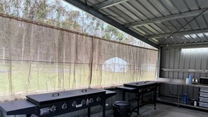 Uma área de cozinha externa com duas churrasqueiras a gás, uma lixeira e uma mesa de metal com micro-ondas.