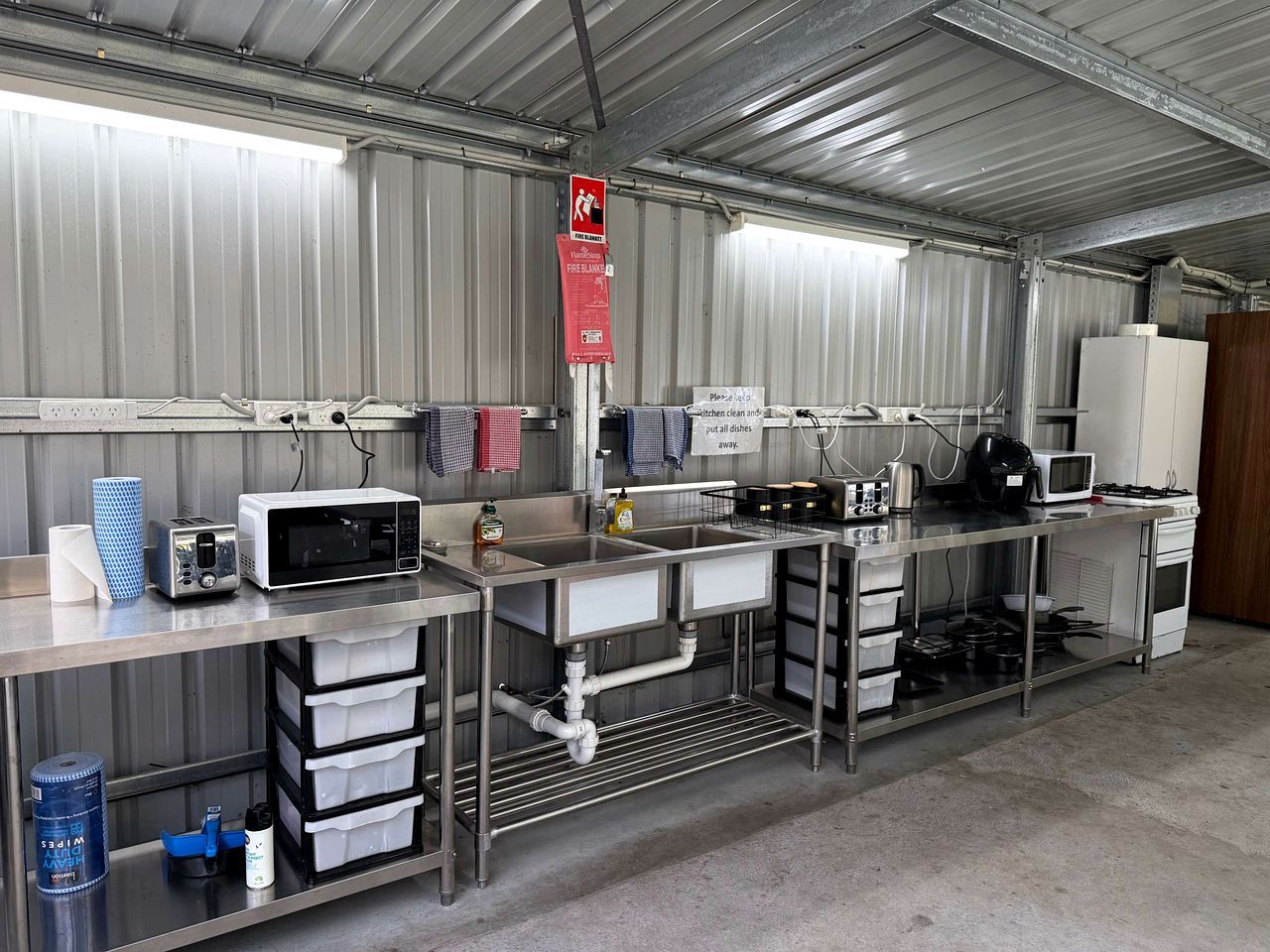 A commercial kitchen with stainless steel appliances, including a microwave, toaster, and sink. There are storage cabinets and a fire blanket sign.