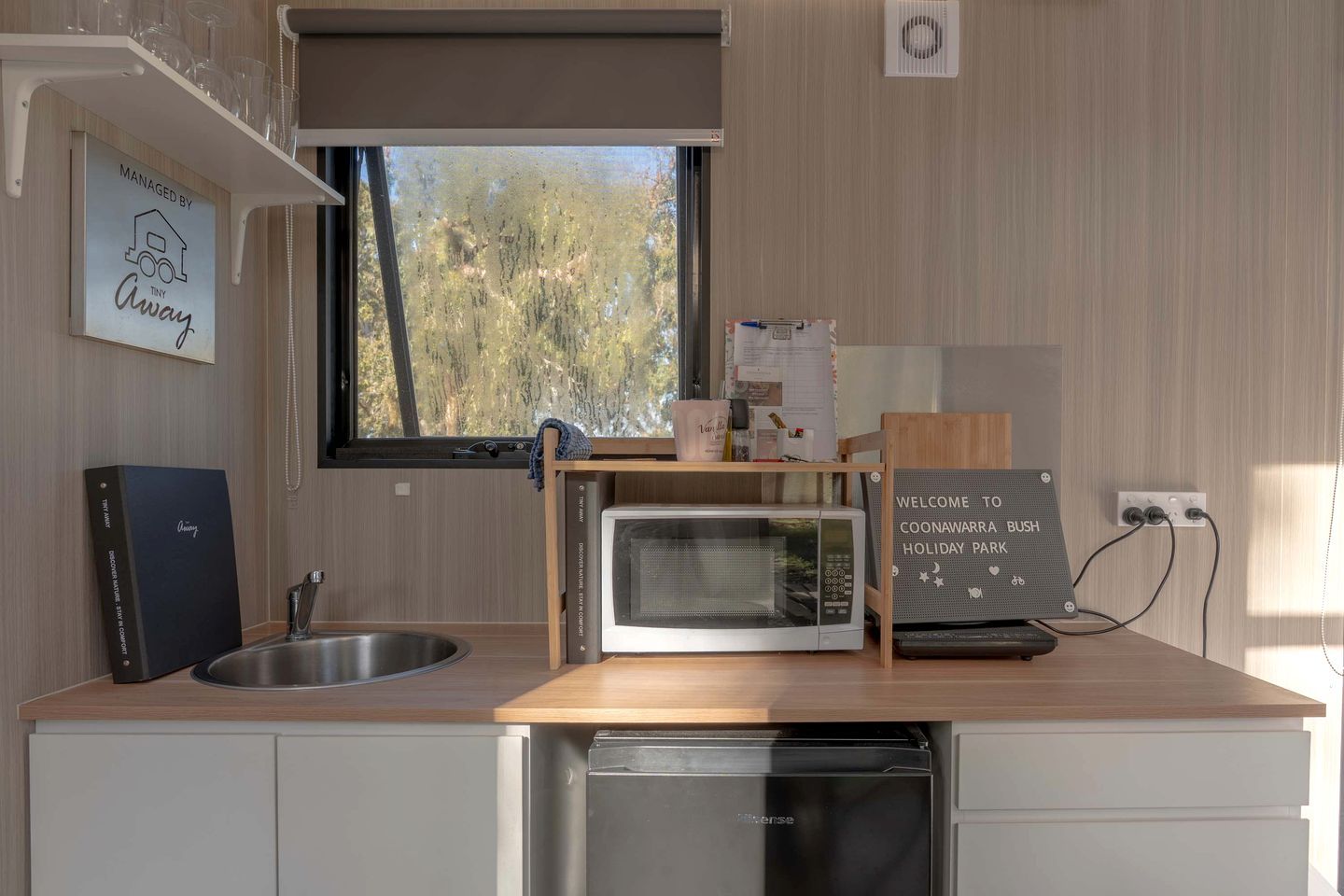A compact kitchen with a sink, microwave, and dishwasher. A welcome sign indicates the location is Coonawarra Bush Holiday Park.