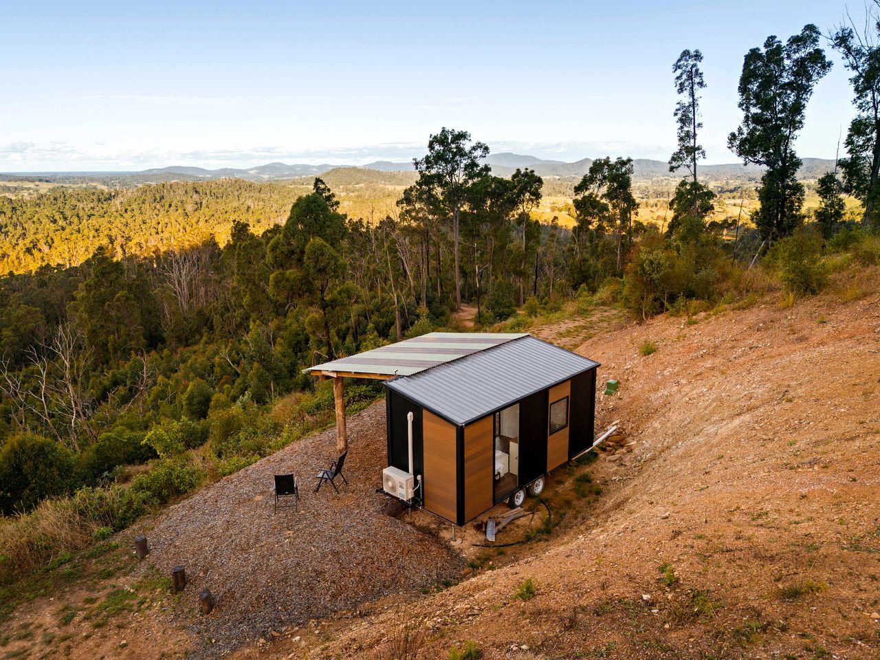 Una cabaña pequeña de un solo piso con techo de metal, ubicada en un sendero de tierra rodeada de árboles y colinas.
