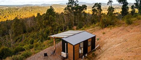 Una cabaña pequeña de un solo piso con techo de metal, ubicada en un sendero de tierra rodeada de árboles y colinas.