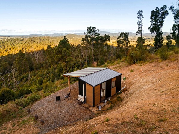 A small, single-story cabin with a metal roof, situated on a dirt path surrounded by trees and hills.