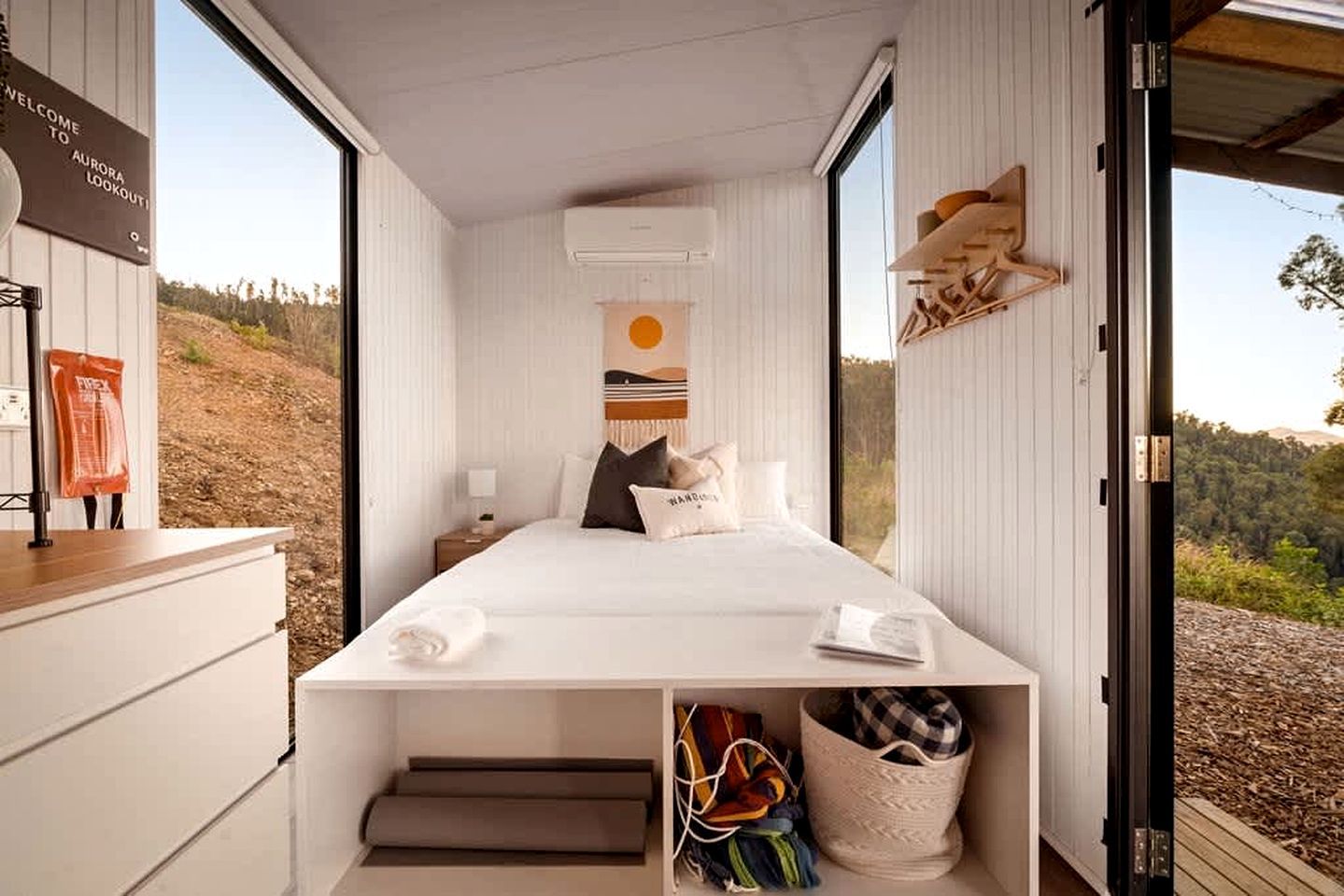 A bedroom with a bed, a nightstand, a wall-mounted shelf, and a window with a view of the outdoors.