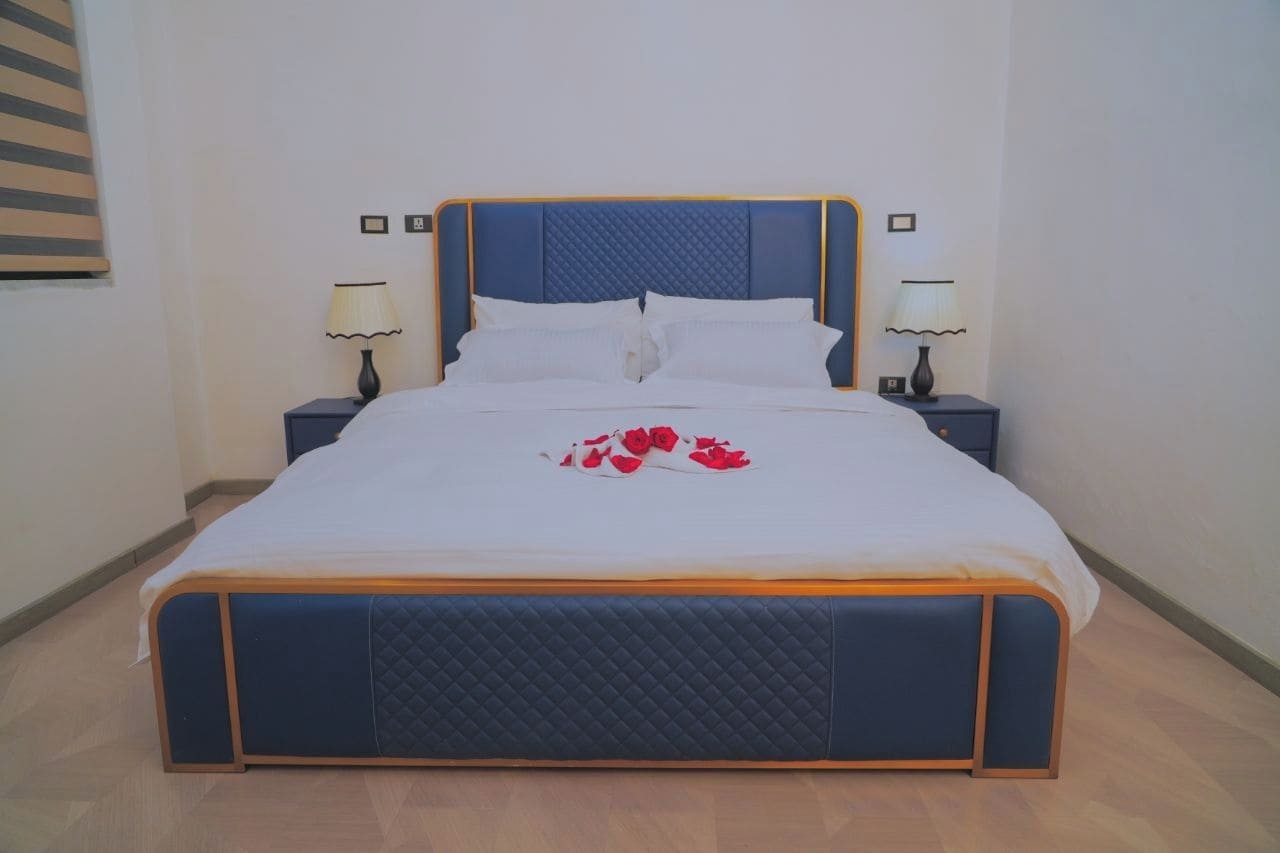 Una cama bien hecha con cabecera, dos mesitas de noche con lámparas, y un arreglo decorativo de rosas rojas sobre la colcha.