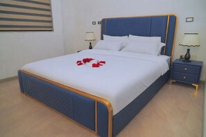 A neatly made bed with a blue headboard and white bedding, a bedside table with a lamp, and a wall-mounted picture frame. - Dooka Apartment (Addis Ababa)
