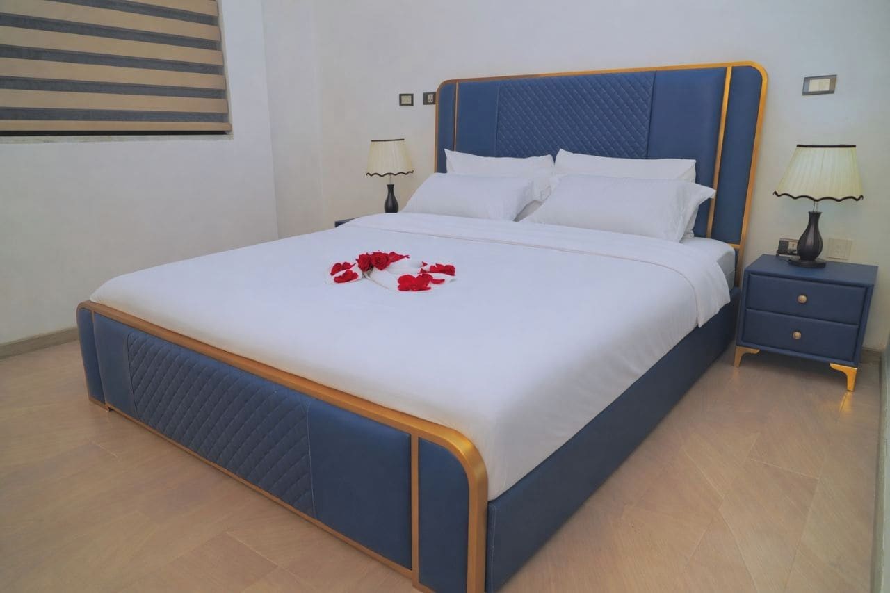 A neatly made bed with a blue headboard and white bedding, a bedside table with a lamp, and a wall-mounted picture frame.