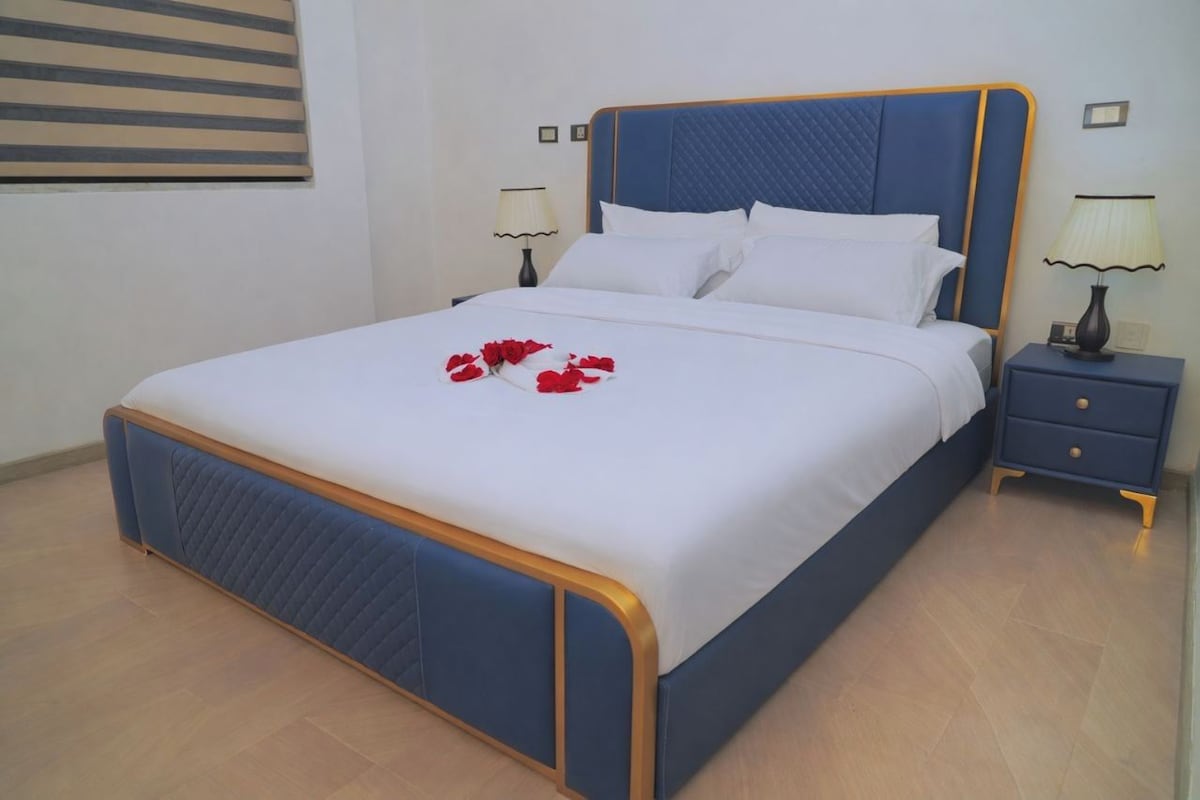 A neatly made bed with a blue headboard and white bedding, a bedside table with a lamp, and a wall-mounted picture frame.