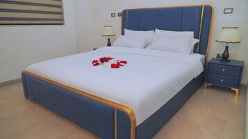 A neatly made bed with a blue headboard and white bedding, a bedside table with a lamp, and a wall-mounted picture frame.