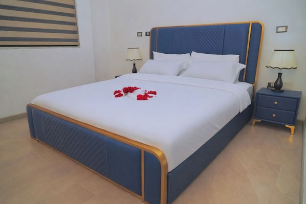 A neatly made bed with a blue headboard and white bedding, a bedside table with a lamp, and a wall-mounted picture frame. - Dooka Apartment (Addis Ababa)