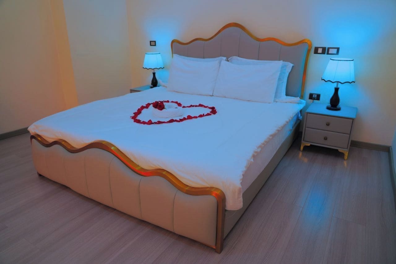 Una cama bien hecha con un adorno con forma de corazón en la colcha, dos mesitas de noche con lámparas y un cabecero de madera.
