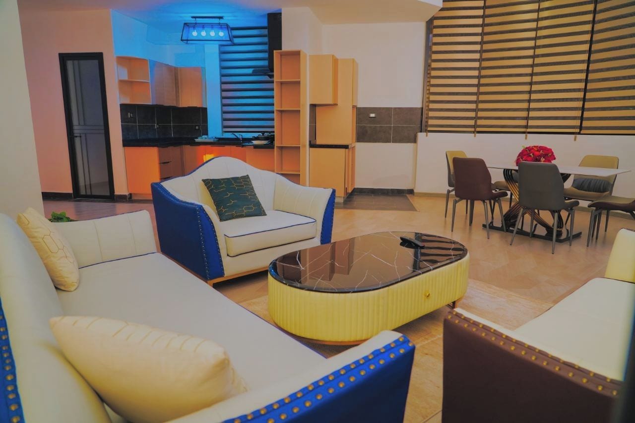 A modern living room with a sofa, armchair, and coffee table. A kitchen with cabinets and a dining area with chairs is visible in the background.