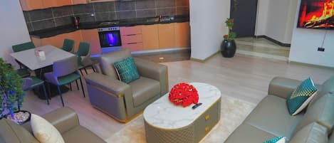 A modern living room with a sofa set, a coffee table with a red floral centerpiece, and a kitchen area with a TV mounted on the wall.