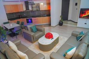 A modern living room with a sofa set, a coffee table with a red floral centerpiece, and a kitchen area with a TV mounted on the wall. - Dooka Apartment (Addis Ababa)