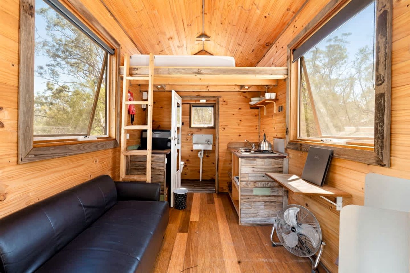 Een compact interieur met houten vloer, een leren zwarte bank, een kitchenette en een ventilator.