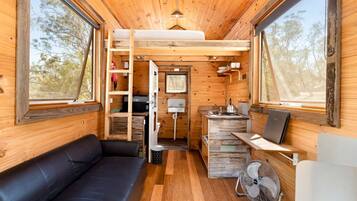 Een compact interieur met houten vloer, een leren zwarte bank, een kitchenette en een ventilator.