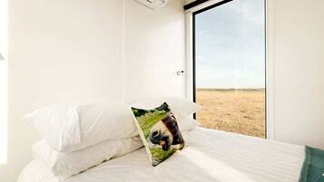 Una cama con ropa blanca y un cojín con la imagen de un panda, una ventana con vistas a un paisaje llano y un equipo de aire acondicionado en la pared.
