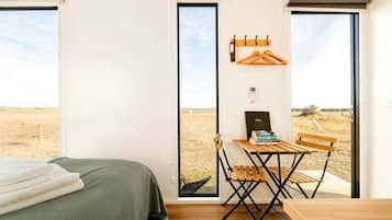 A room with a bed, a desk with a chair, a window with a view of a field, and a coat rack with hangers.