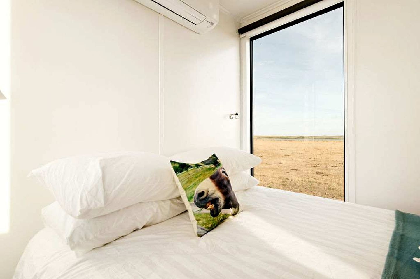 A bed with white bedding and a pillow with a panda image, a window with a view of a flat landscape, and an air conditioning unit on the wall.